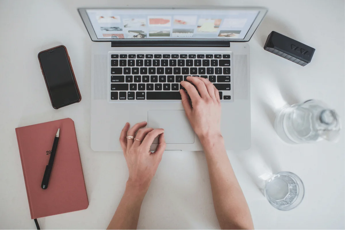 Hands typing on a laptop surrounded by a red notebook, smartphone, and a glass of water.