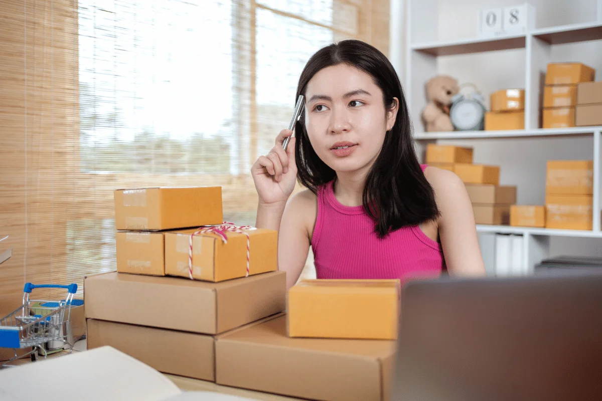 Female entrepreneur in a pink top planning e-commerce orders, surrounded by shipping boxes in a home office.