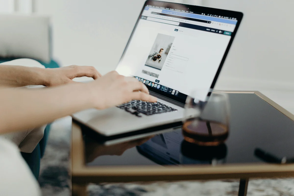 Person typing on a laptop at a glass table, with a drink in the foreground and a minimalist home interior.