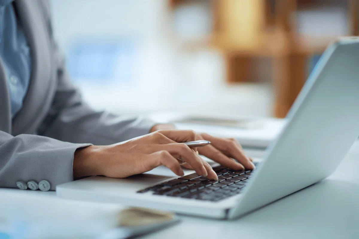 Professional woman in a gray suit typing on a laptop, with a pen in hand, at a modern office desk.