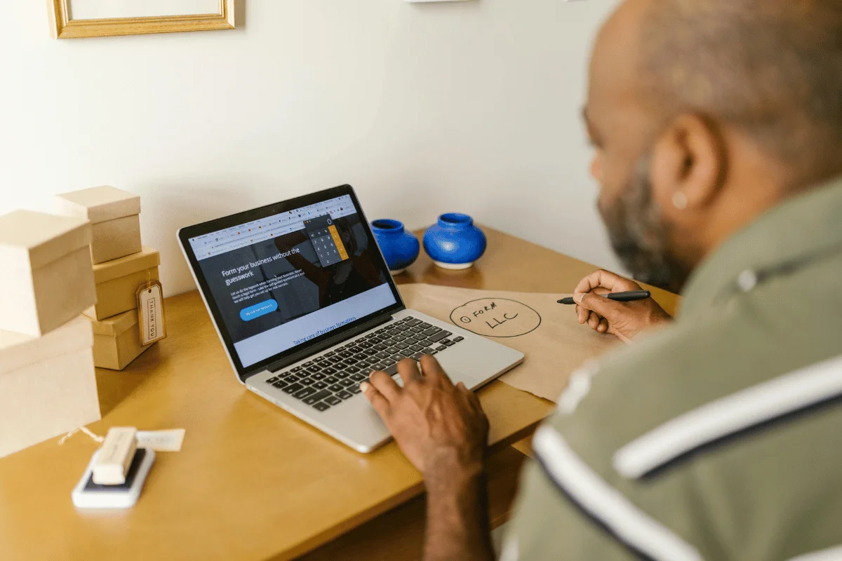 Man working on a laptop at a desk, writing "Form LLC" on a paper, with small boxes and blue vases in the background.