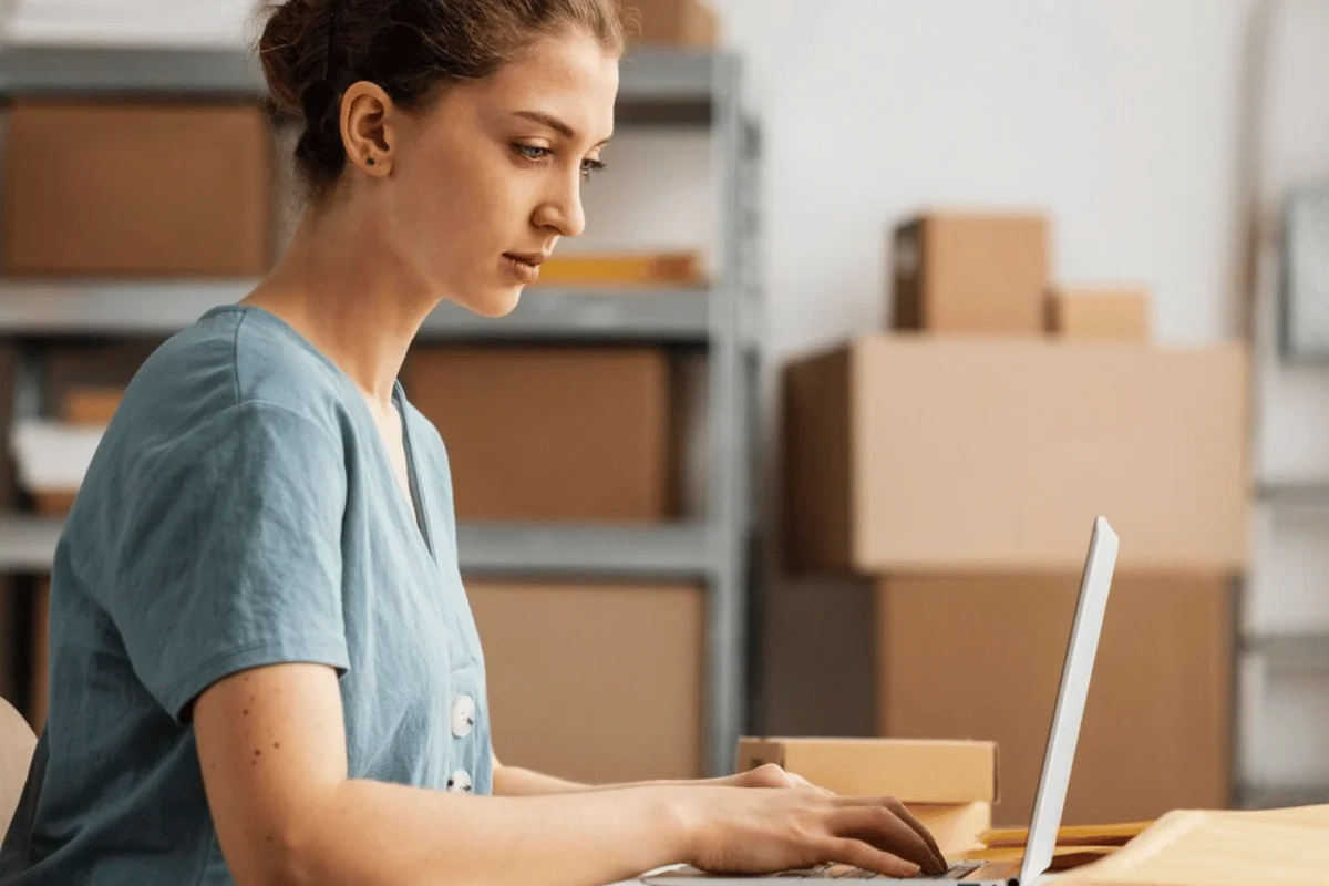 Female office worker using a laptop with shelves and cardboard boxes in the background.