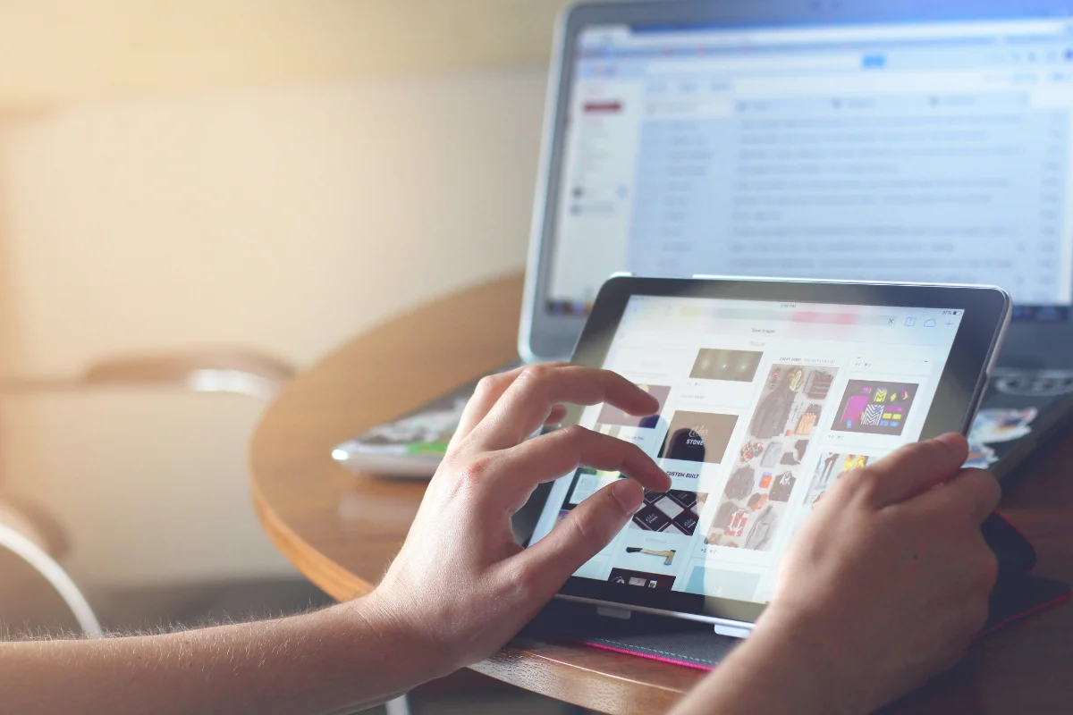 Person using a tablet with a laptop in the background on a wooden table.