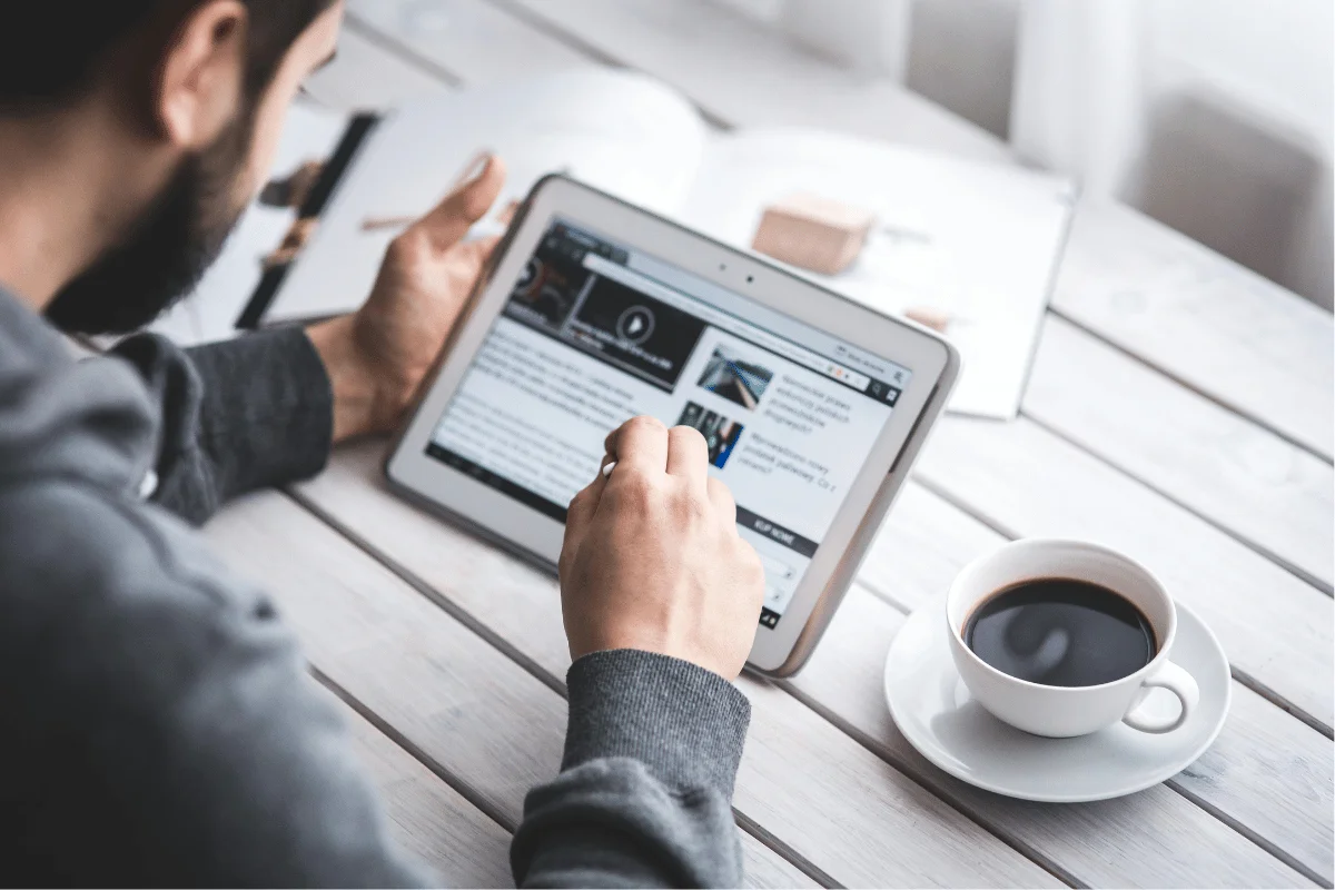 Person using a tablet to browse the internet while drinking coffee at a wooden table.