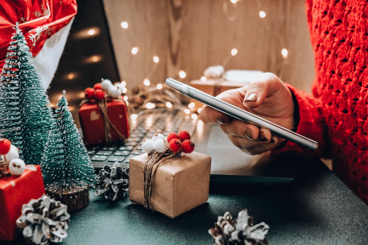 Person browsing on tablet amidst Christmas decor and gifts, with warm lights.