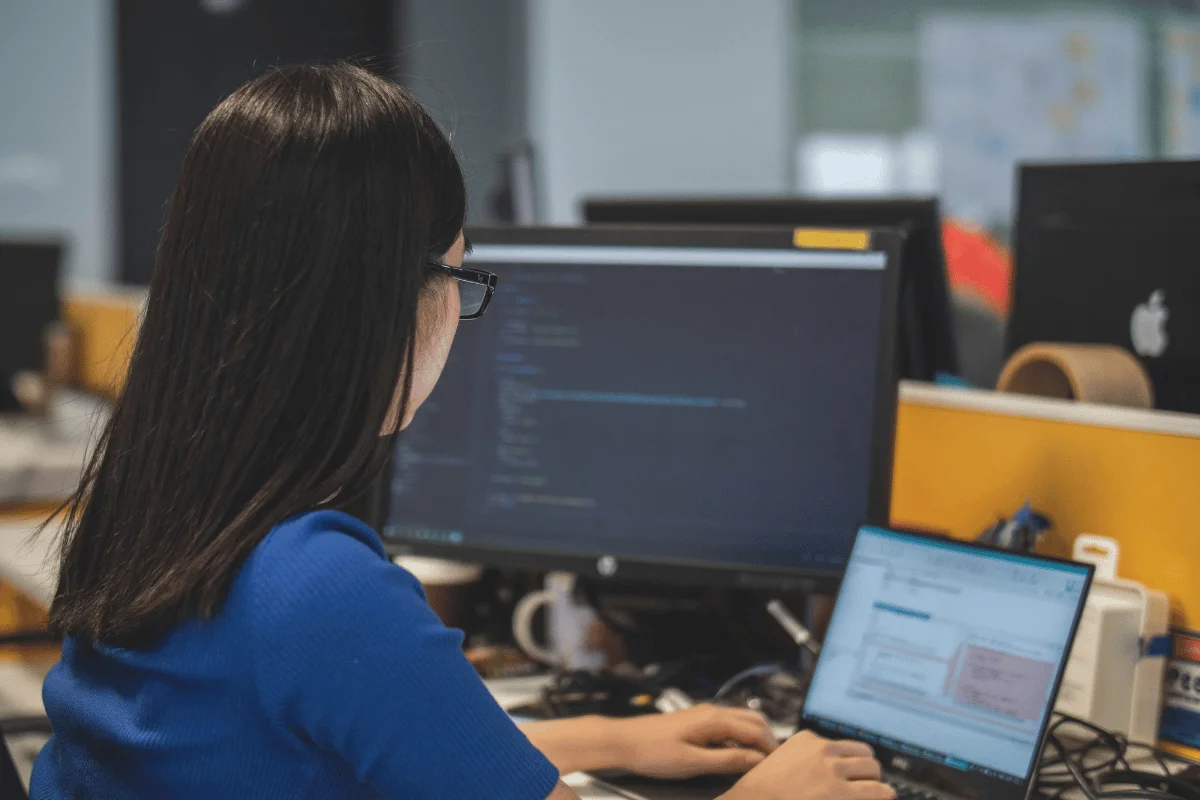 A woman wearing glasses, seated at a desk, types on a laptop with code displayed on two computer monitors behind her.