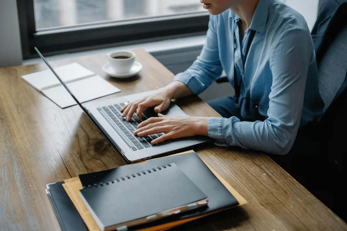Person working on a laptop with notepads and a coffee cup on a wooden table.