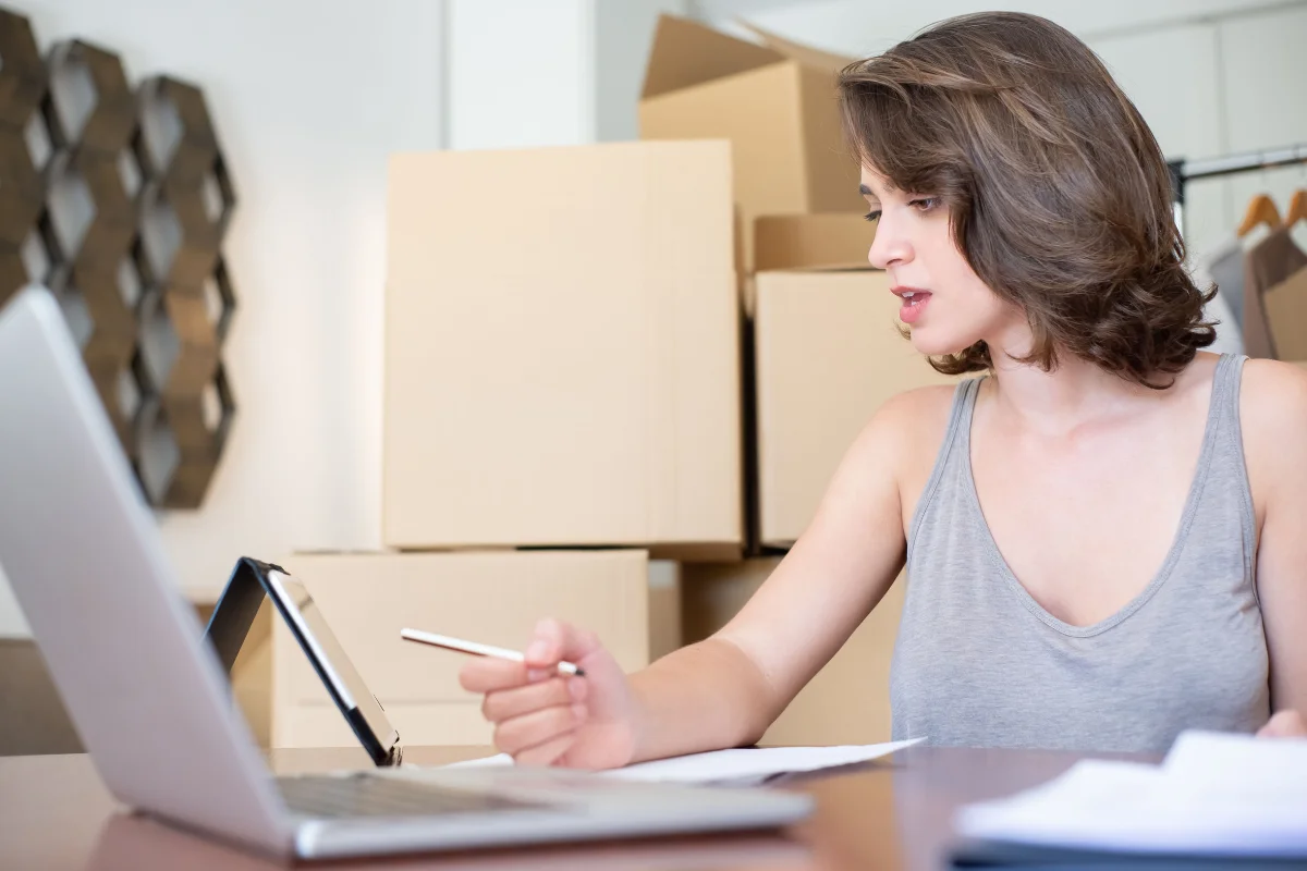 Focused woman writing beside an open laptop at a desk, surrounded by stacked cardboard boxes, suggesting home office work or moving.
