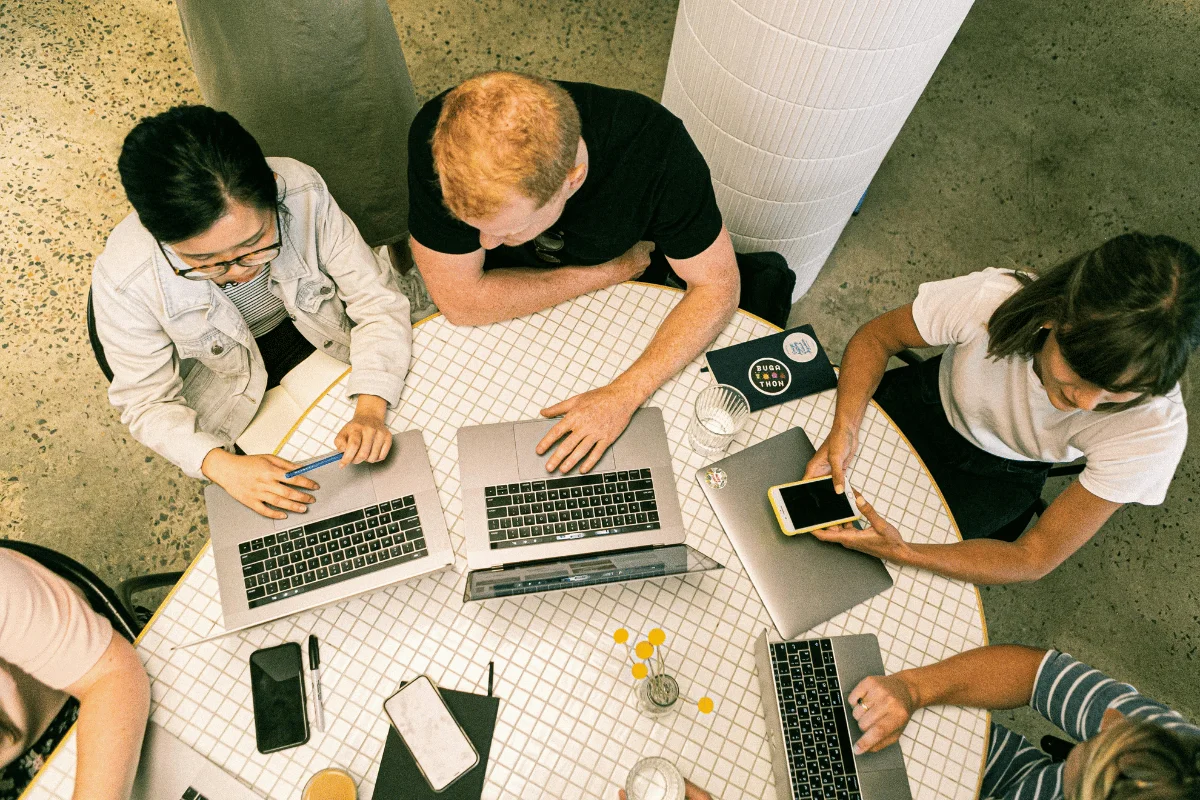 A group of five people collaborates around a round table with laptops and phones. The setting is informal, conveying teamwork and creativity.