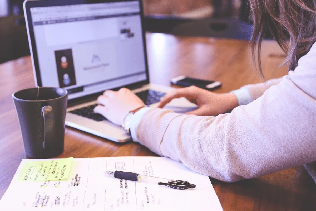 Close-up of a person working on a laptop with a cup and pens on a desk.