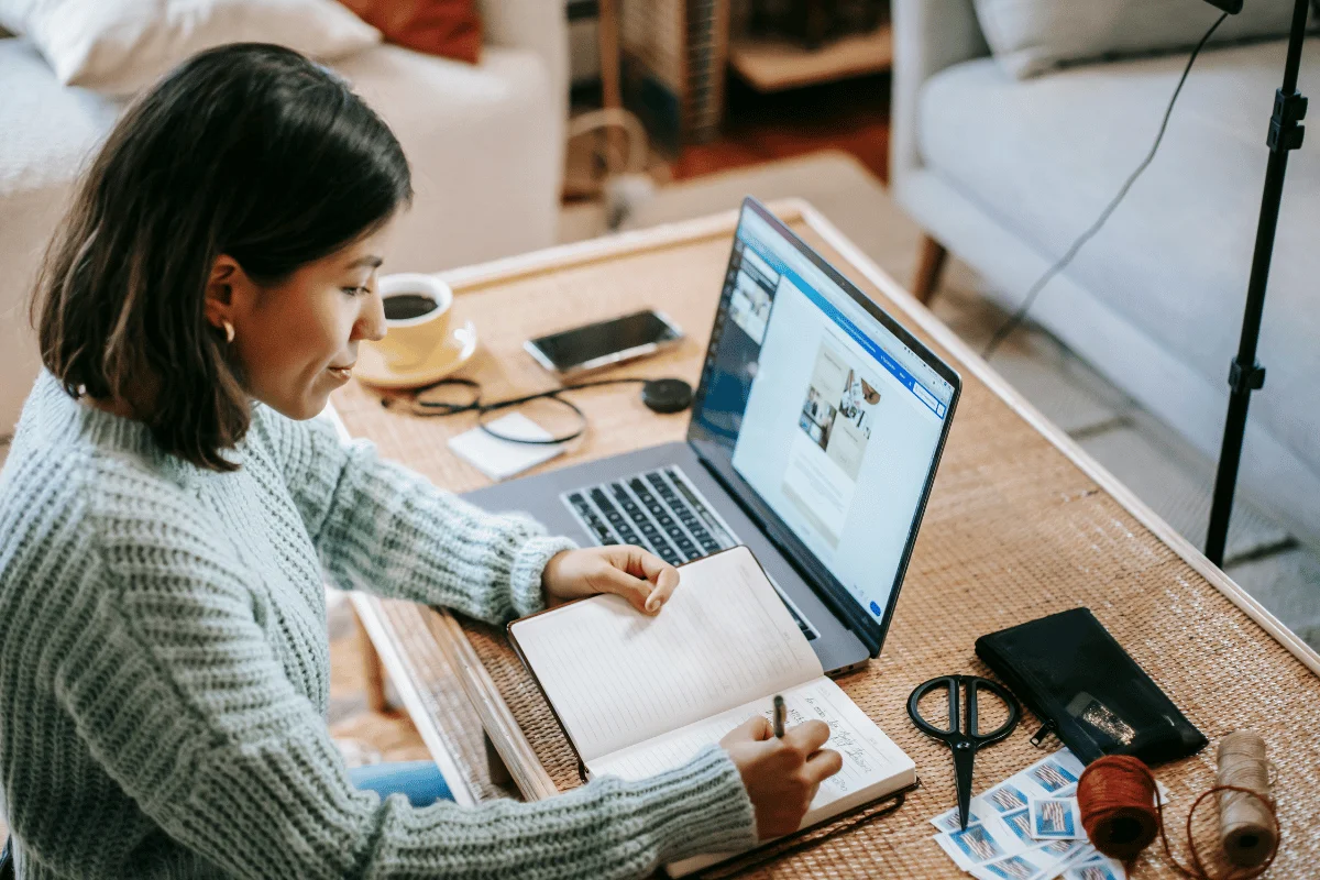 Woman in a cozy sweater writing in a notebook at a desk, with a laptop displaying a webpage, and crafting materials around her, creating a focused atmosphere.
