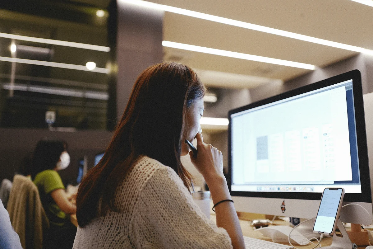 A person in a cozy sweater sits at a computer, focused on design work, with a smartphone displayed nearby.
