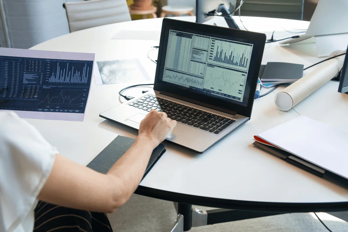 A person analyzing financial data on a laptop, surrounded by charts, notebooks, and office supplies on a round table.