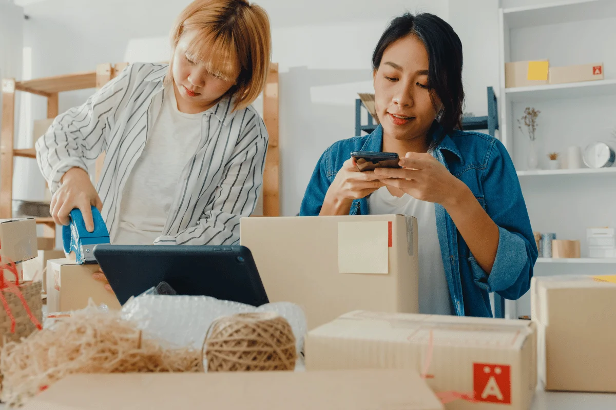 Two people are packing boxes in a workspace, one using tape and the other checking their phone, surrounded by packing materials.