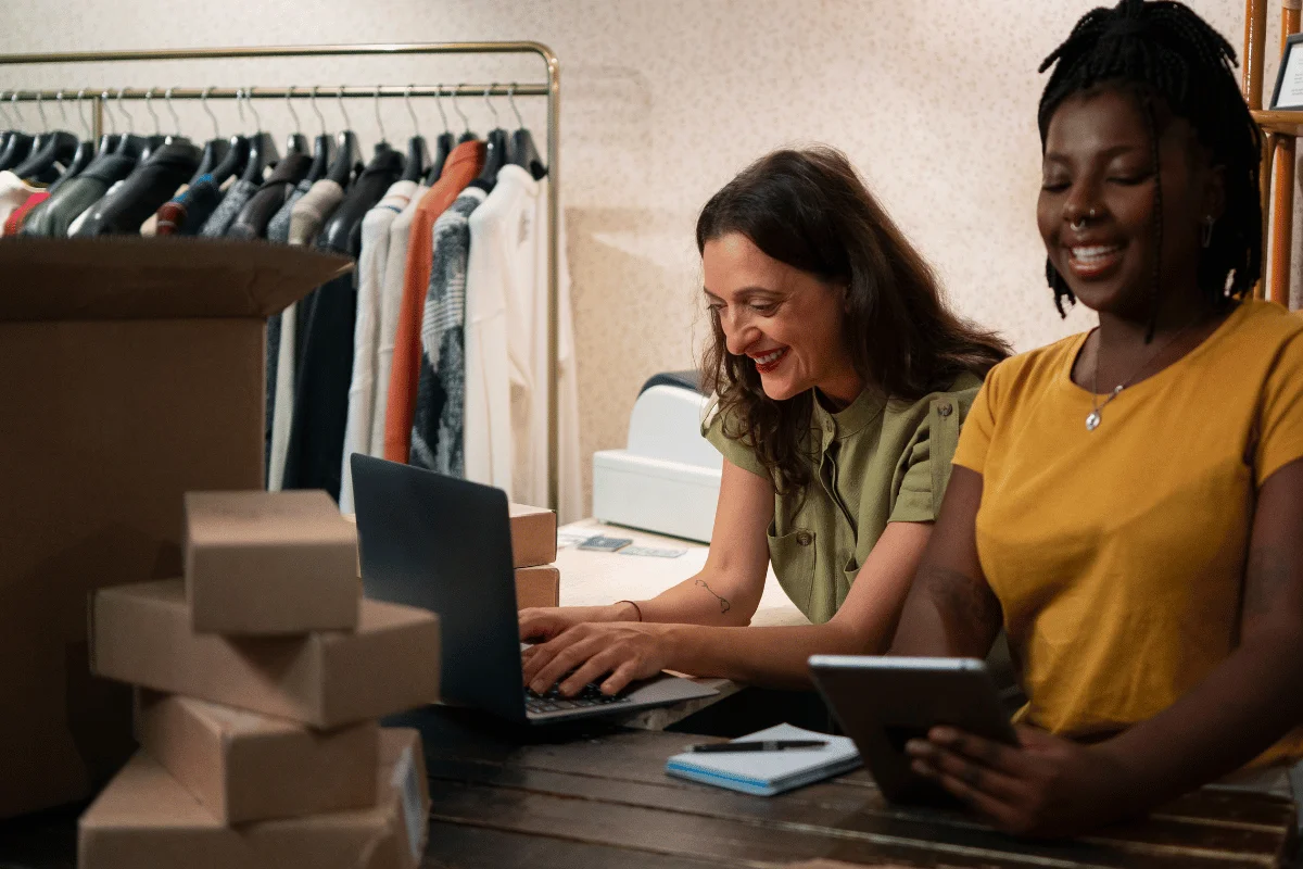 Two women are joyfully working in a clothing store. One types on a laptop surrounded by boxes, while the other holds a tablet.