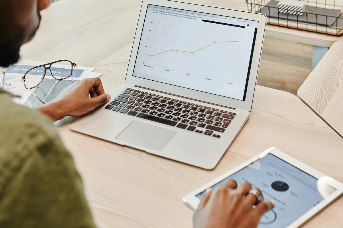 A person views statistical graphs on a laptop and a tablet, analyzing data with a focused expression in a bright workspace.