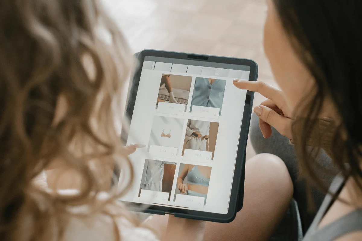 Two women sit closely, browsing clothing options on a tablet.
